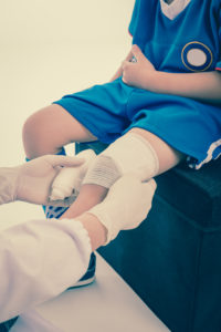Sports injury. Youth athlete child in blue uniform. Doctor perform first aid knee injury by bandage. Shoot in studio. Cream tones.